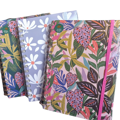A close-up view of three A5 wire-bound notebooks with assorted vibrant cover prints, including a tropical leaf pattern and a pastel daisy pattern.
