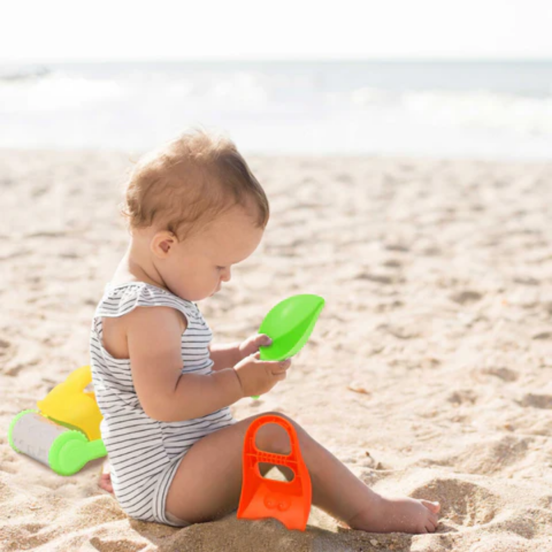 Toddler sitting on a sandy beach playing with the green Tookyland scoop and orange sand tool.